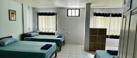 Une chambre avec quatre lits simples, une armoire, une fenêtre avec des rideaux et un sol carrelé.