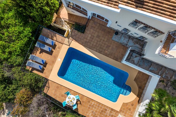 An aerial view of a house with a swimming pool, a patio with a table and chairs, and a fenced garden area.