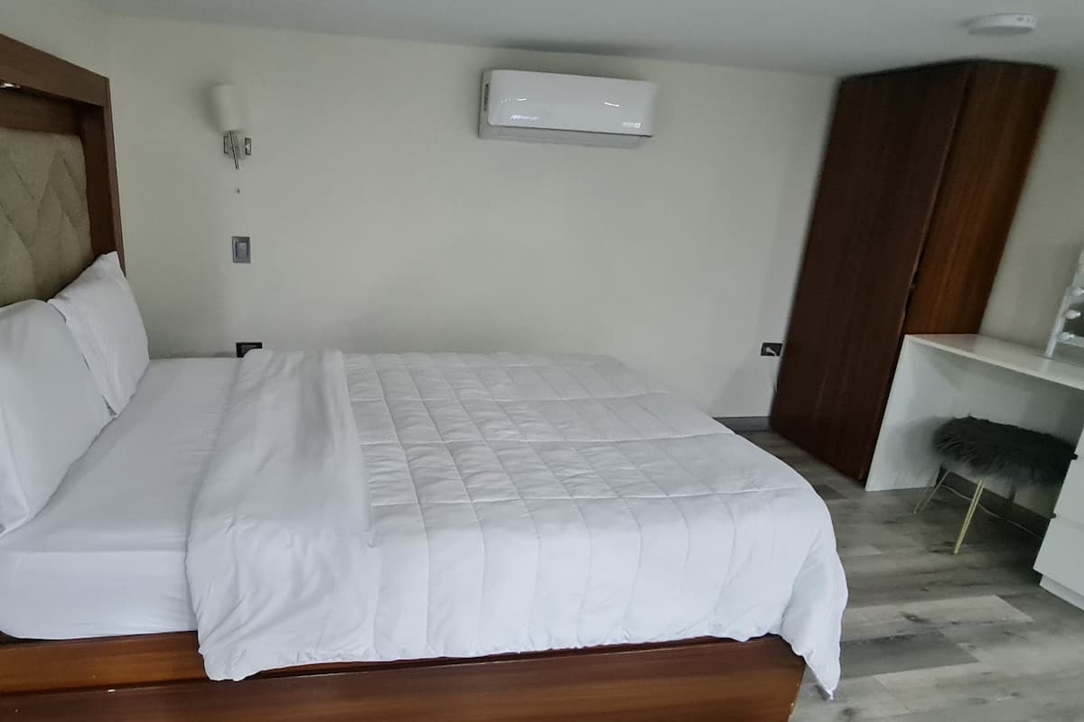 A neatly made bed with a wooden headboard, a white bedside table, a mirror with lights, and a white wardrobe.