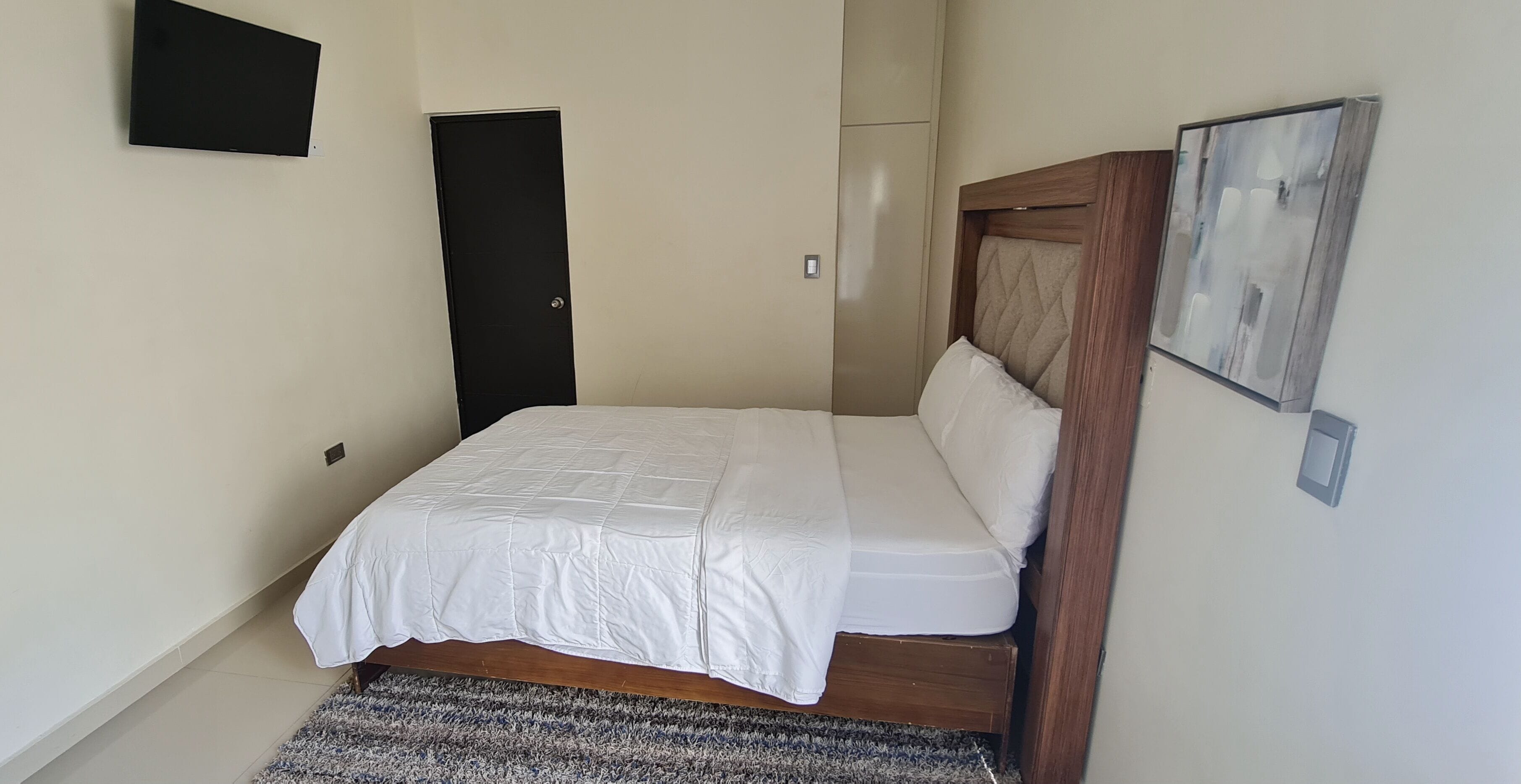 A bedroom with a bed, a door, a wall-mounted light, and a framed picture.