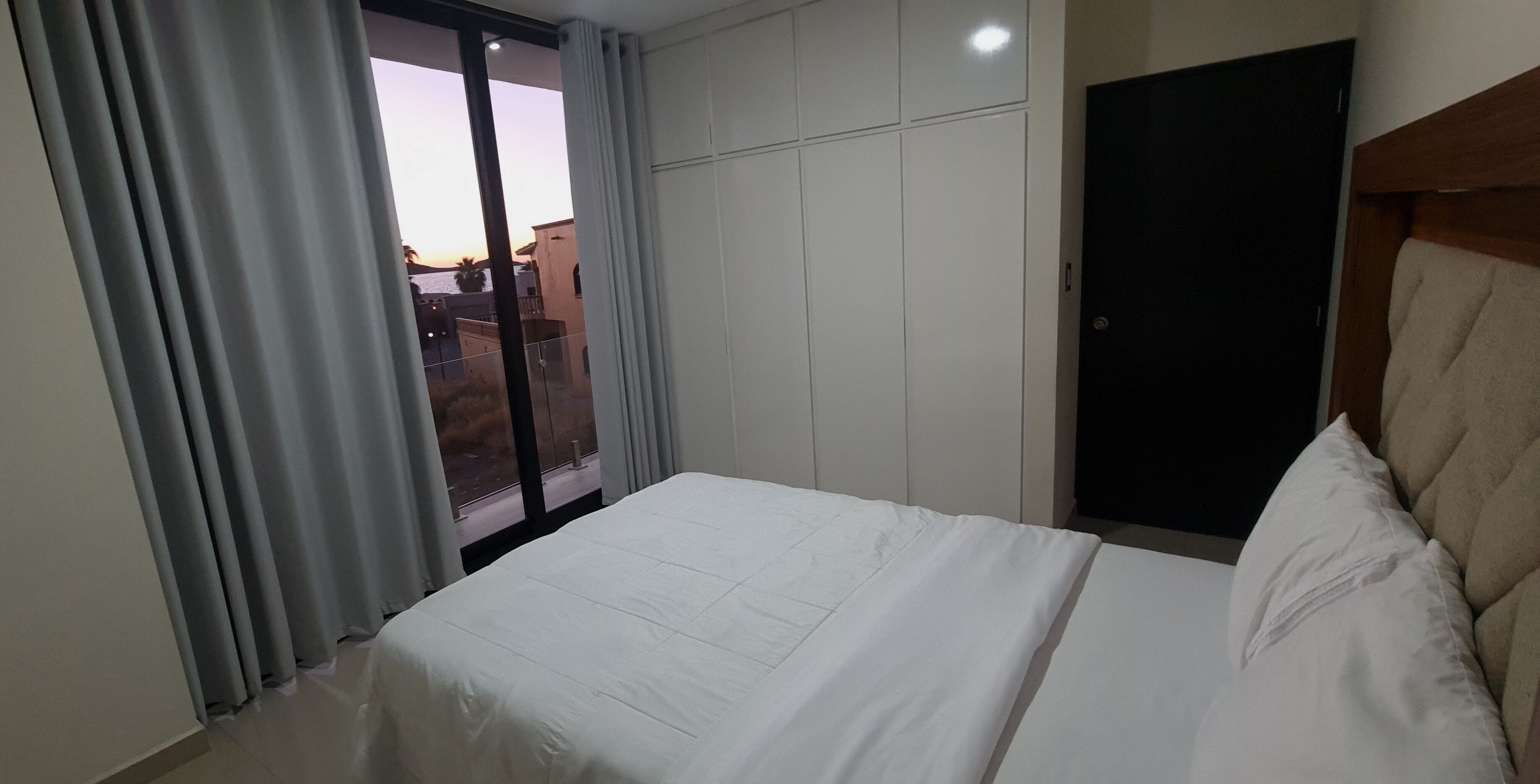 Una habitación de hotel con una cama, un ventanal corredero al exterior y una puerta oscura.