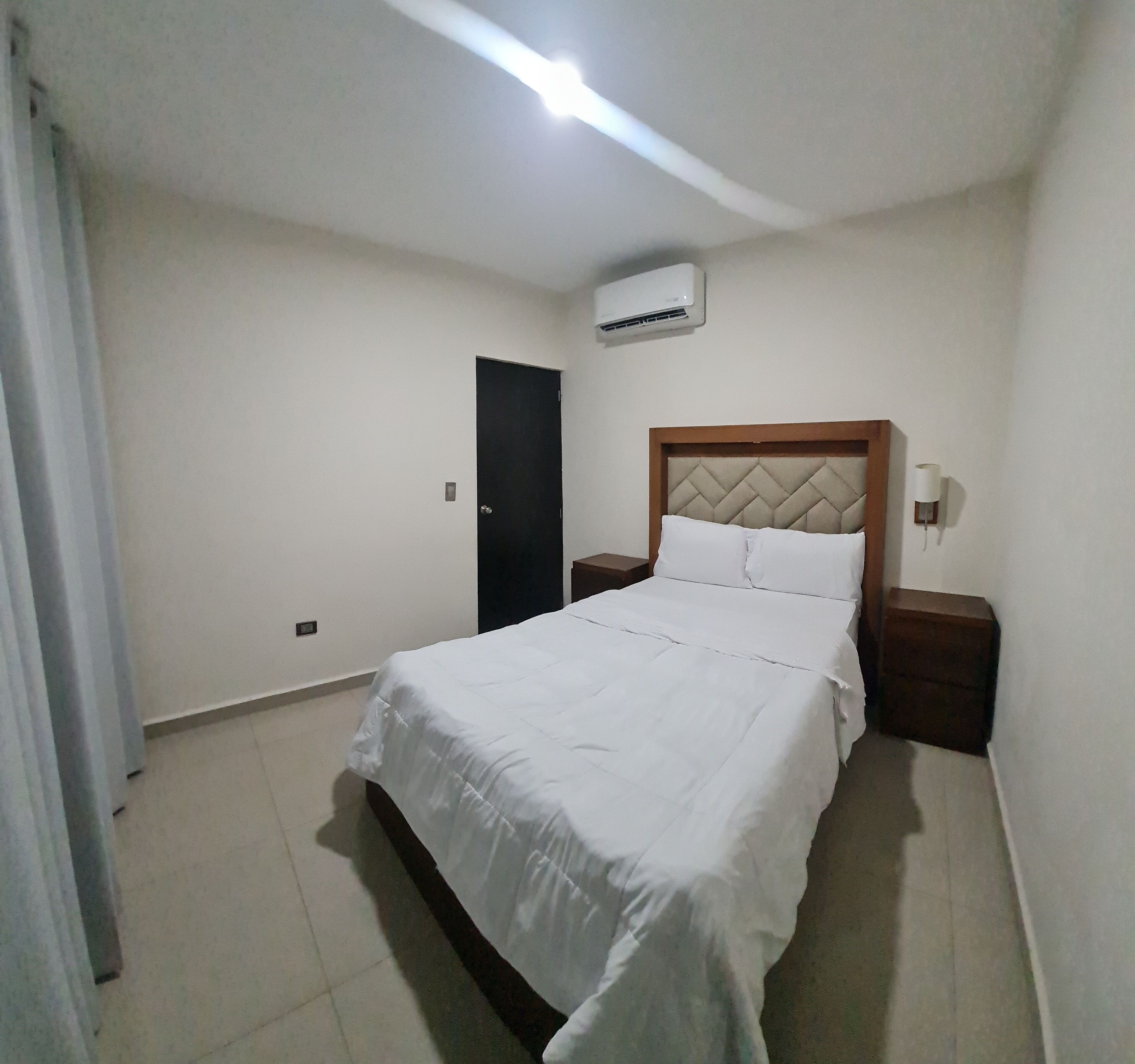 A bedroom with a bed, a nightstand, a wall-mounted air conditioner, and a door.