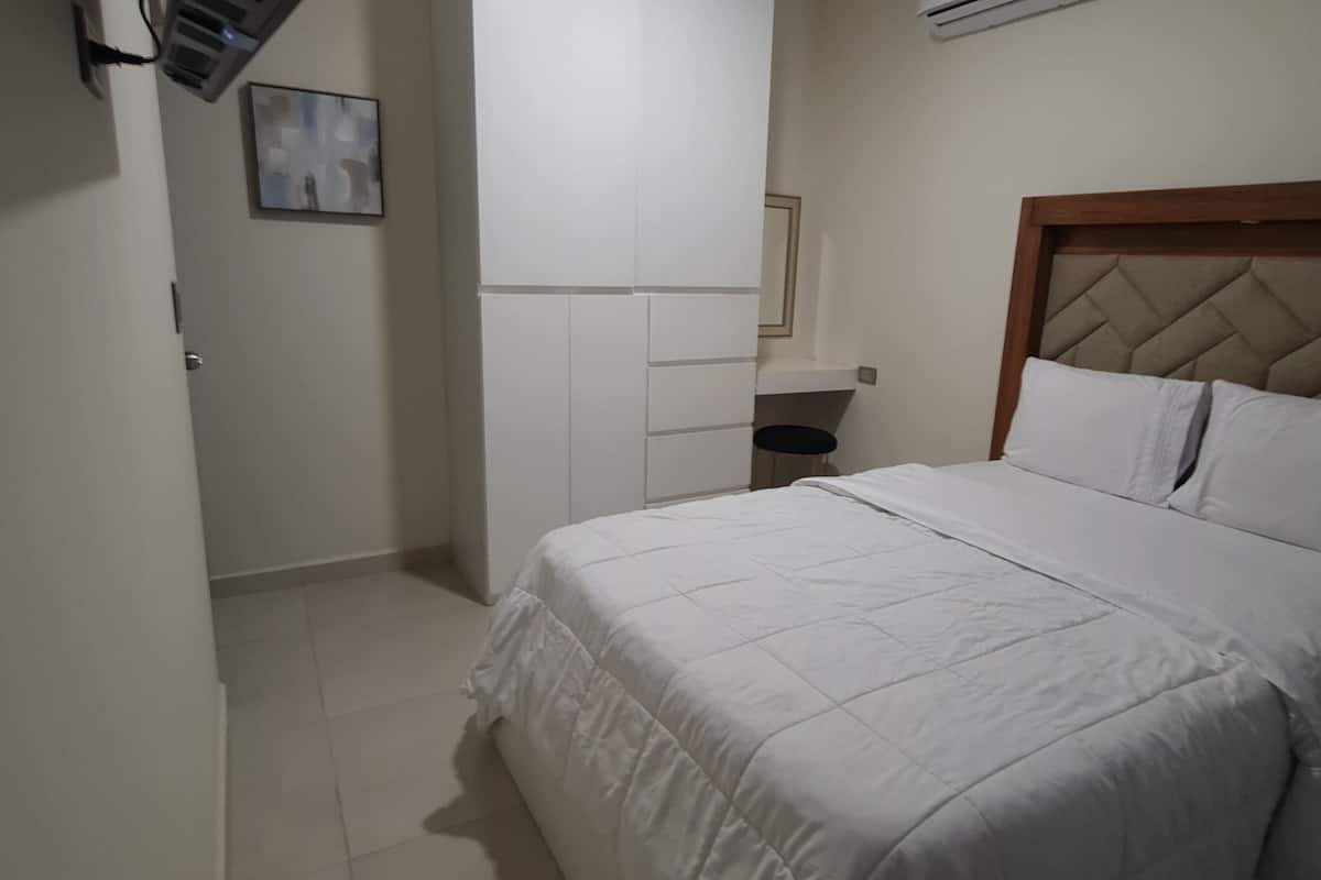 A bedroom with a bed, a bedside table, a mirror, and a wall-mounted air conditioner.