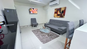 Ein modernes Wohnzimmer mit einem grauen Sofa, einem Glastisch, einem grauen Sessel und einem an der Wand montierten Fernseher.