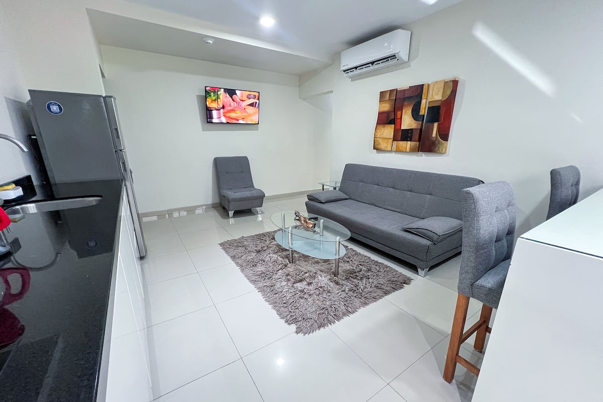 A modern living room with a grey sofa, a glass coffee table, a grey armchair, and a TV mounted on the wall.