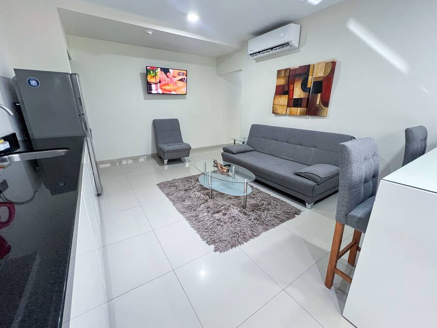 A modern living room with a grey sofa, a glass coffee table, a grey armchair, and a TV mounted on the wall.