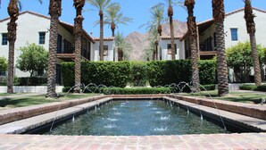 A courtyard with a central water feature, surrounded by palm trees and a building with multiple windows. - Desert Daydream A | 1BR Legacy Villa (La Quinta)