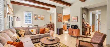 A spacious living room with a sofa set, a coffee table, and a chair. There is a kitchen in the background with wooden cabinets and a dining area.