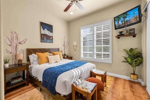 A bedroom with a bed, a nightstand, a chair, a window with blinds, a ceiling fan, and a wall-mounted TV. - Desert Daydream A | 1BR Legacy Villa (La Quinta)