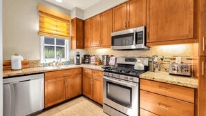A modern kitchen with wooden cabinets, stainless steel appliances, and a window with blinds. - Desert Daydream A | 1BR Legacy Villa (La Quinta)
