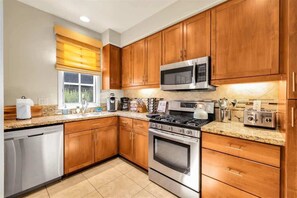 A modern kitchen with wooden cabinets, stainless steel appliances, and a window with blinds. - Desert Daydream A | 1BR Legacy Villa (La Quinta)