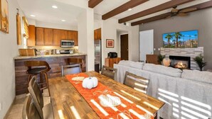 A modern kitchen with wooden cabinets, a dining area with a table set for four, and a living room with a fireplace. - Desert Daydream A | 1BR Legacy Villa (La Quinta)