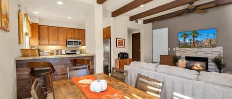 Una cocina moderna con armarios de madera, un comedor con una mesa preparada para cuatro personas y una sala de estar con chimenea.