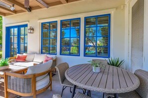 Une terrasse couverte, aménagée avec des meubles en osier, une table ronde et de grandes fenêtres dotées de volets bleus.
