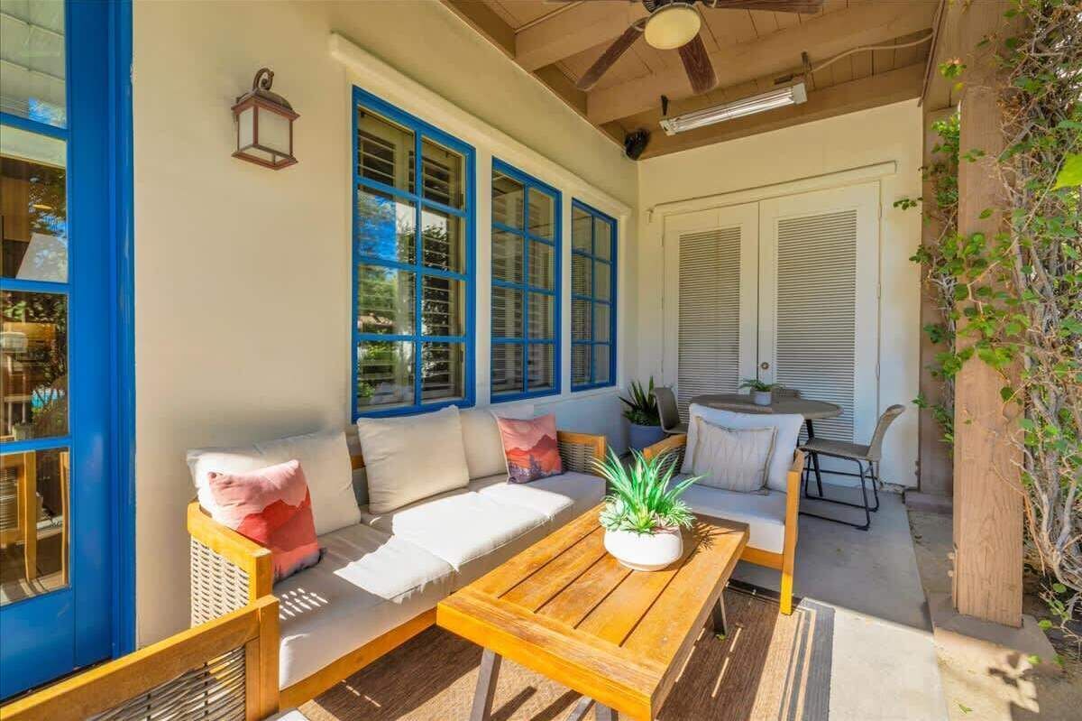 A sunlit patio with a wooden table, cushioned seating, and large windows with blue frames.