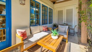 A sunlit patio with a wooden table, cushioned seating, and large windows with blue frames. - Desert Daydream A | 1BR Legacy Villa (La Quinta)