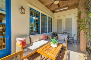 A sunlit patio with a wooden table, cushioned seating, and large windows with blue frames.