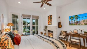 A bedroom with a bed, a desk, a guitar, a fireplace, and a painting of palm trees. - Desert Daydream B | Studio Legacy Villa (La Quinta)