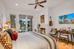 A bedroom with a bed, a desk, a guitar, a fireplace, and a painting of palm trees. - Desert Daydream B | Studio Legacy Villa (La Quinta)