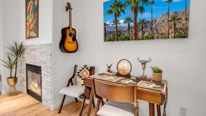 A living room with a fireplace, a guitar, a painting of palm trees, and a desk with a mirror and decorative items. - Desert Daydream B | Studio Legacy Villa (La Quinta)