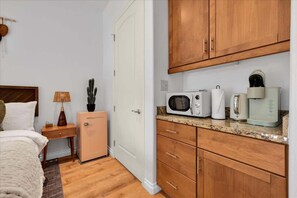 A compact bedroom with a bed, bedside table, lamp, and a small refrigerator. Adjacent is a kitchenette with a microwave, toaster, and coffee maker.