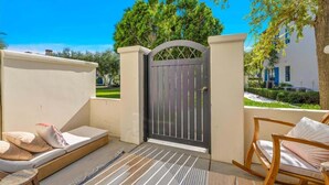 A balcony with a mattress and pillows, a wooden rocking chair, and a gated entrance with a view of a garden and a building. - Desert Daydream C | Studio Legacy Villa (La Quinta)