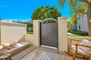A balcony with a mattress and pillows, a wooden rocking chair, and a gated entrance with a view of a garden and a building.