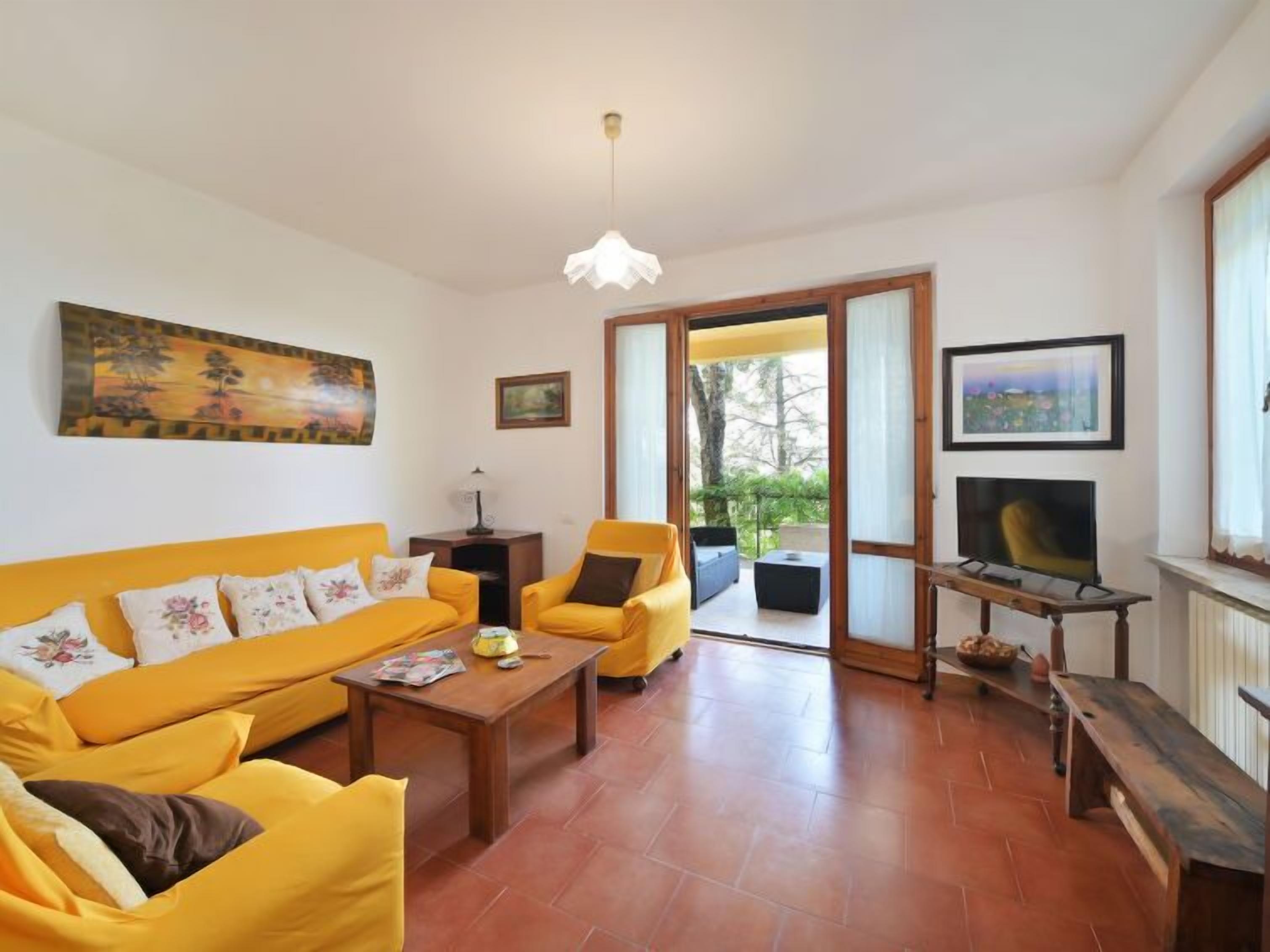 Una sala de estar con un sofá amarillo, una mesa de centro y un televisor. Hay un cuadro en la pared y una puerta que da al exterior.