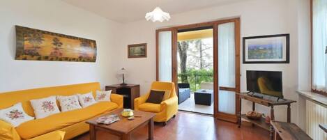 Una sala de estar con un sofá amarillo, una mesa de centro y un televisor. Hay un cuadro en la pared y una puerta que da al exterior.