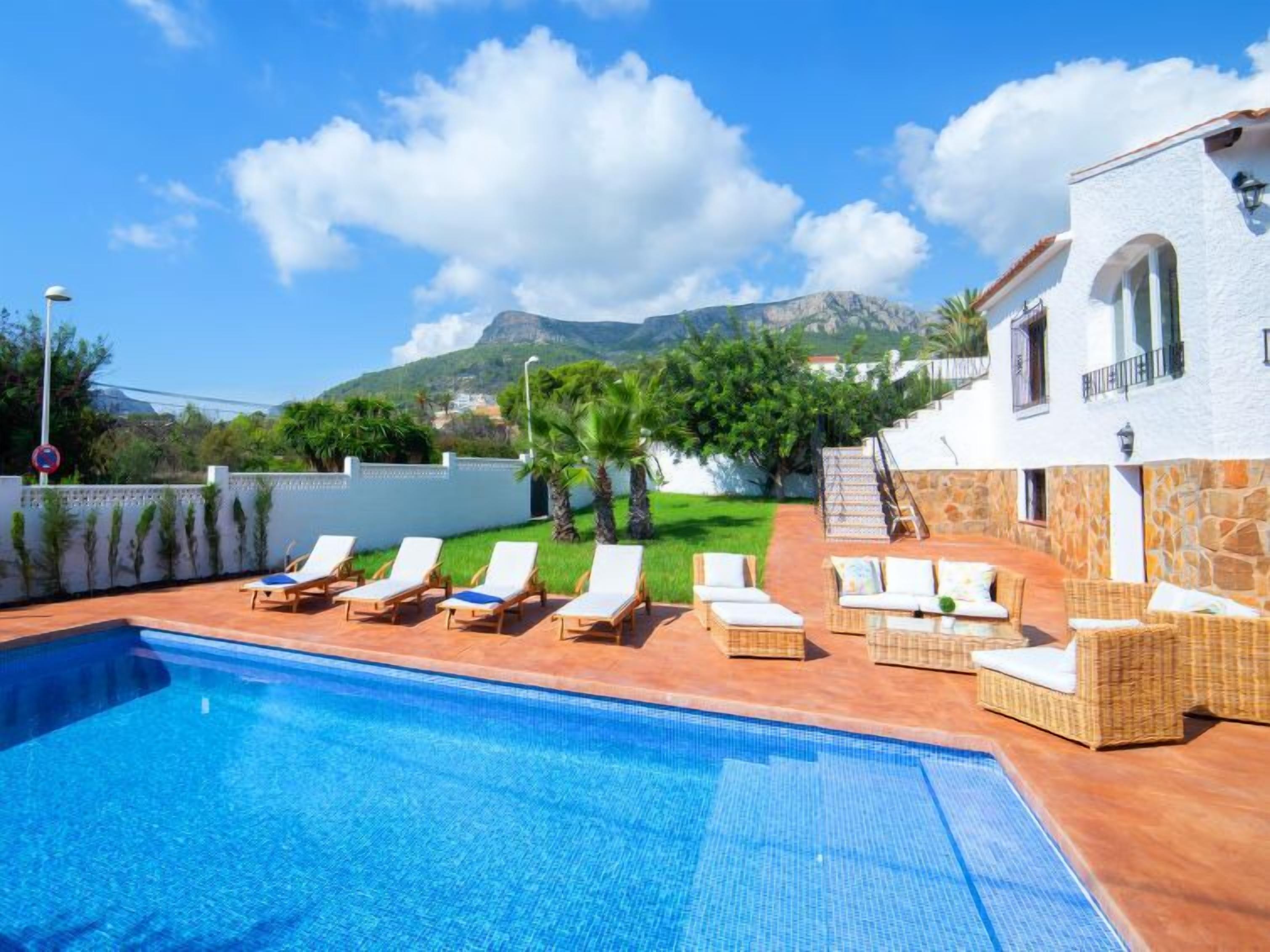 A swimming pool with lounge chairs, a white building with stone walls, and a mountainous backdrop.