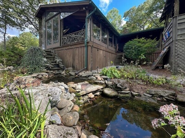 A wooden cabin with a pond, surrounded by rocks and greenery.