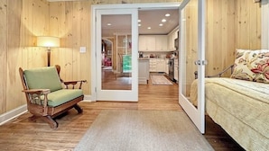 A cozy room with a wooden floor, a green armchair, a bed with a floral pillow, and a kitchen visible through an open door. - Walk to Dinner. Walk to Coffee. Walk to Everything | Wilson Cottage (Highlands)