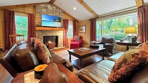 A cozy living room with leather sofas, a fireplace, a television, and large windows offering a view of greenery. - Walk to Dinner. Walk to Coffee. Walk to Everything | Wilson Cottage (Highlands)