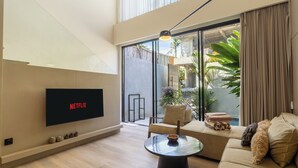 A modern living room with a flat-screen TV displaying 'NETFLIX', a sofa, a coffee table, and a sliding glass door leading to a balcony with plants. - Oasis Villa 9 by Betterplace (Canggu)