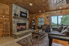 A cozy cabin interior with a stone fireplace, wooden walls, a leather sofa, and a large window offering a view. - Sunrise Overlook | Blue Ridge, GA (Blue Ridge)