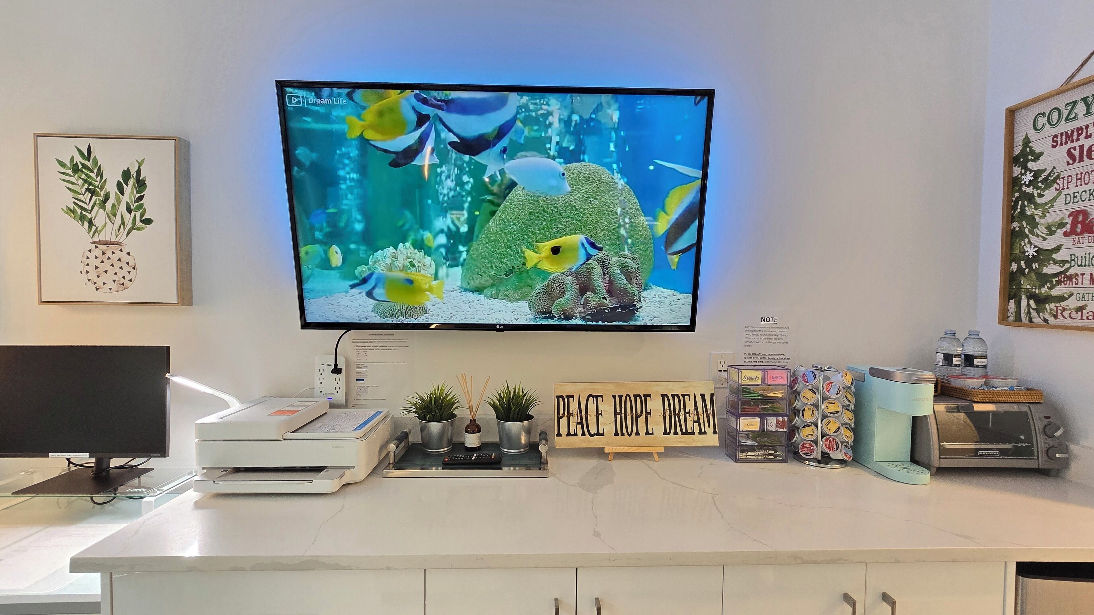 Un espacio de trabajo en casa con un monitor de computadora que muestra un acuario, una impresora, un cafetera y un letrero que dice 'Peace Hope Dream'.
