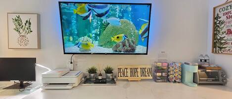 Un espacio de trabajo en casa con un monitor de computadora que muestra un acuario, una impresora, un cafetera y un letrero que dice 'Peace Hope Dream'.