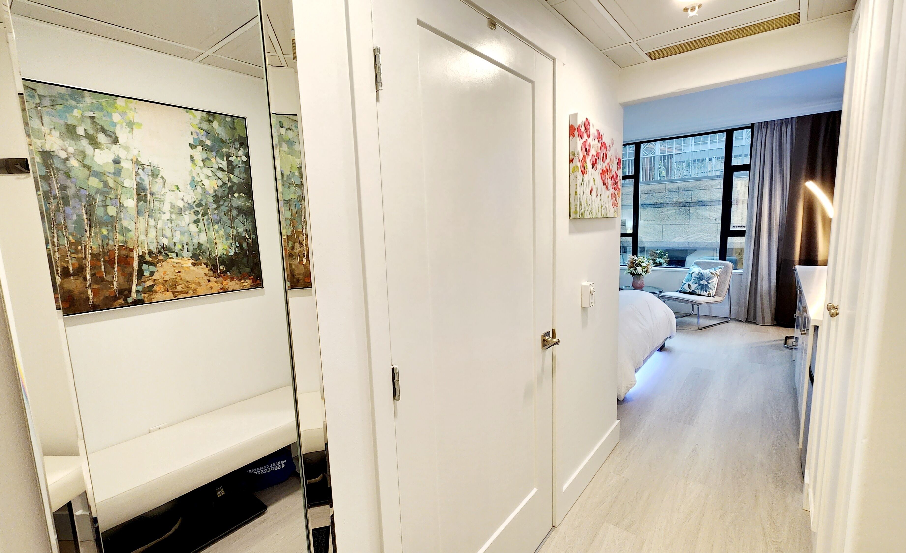 Una habitación de hotel moderna con un ventanal grande, una cama, un área de descanso con una silla y una mesa, y obras de arte en las paredes.