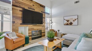 A modern living room with a large flat-screen TV mounted on a wooden wall, a fireplace, comfortable seating, and a large plant centerpiece. - Palmas de Indio: Pool, Private, Walk to Festivals (Indio)