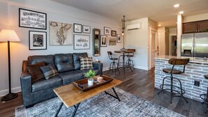 A living room with a sofa, coffee table, and a dining area with a table and chairs. There is a kitchen area with a refrigerator and a bar counter with stools. - Modern 1BR Flat: Free Parking, AC, Getaway Ready! (Lynchburg)