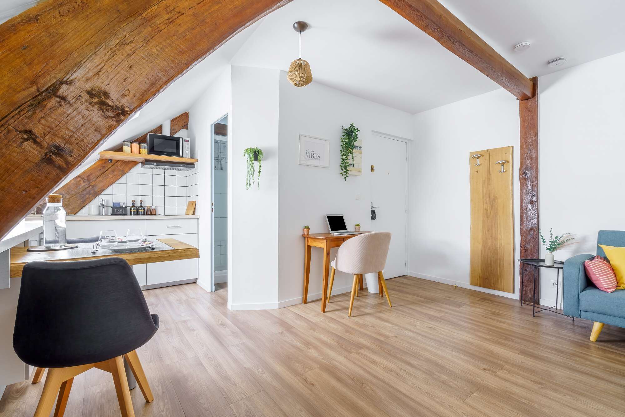 A modern loft apartment with exposed wooden beams, a kitchen with a microwave, a dining area with a table and chairs, and a living room with a sofa and a blue armchair.
