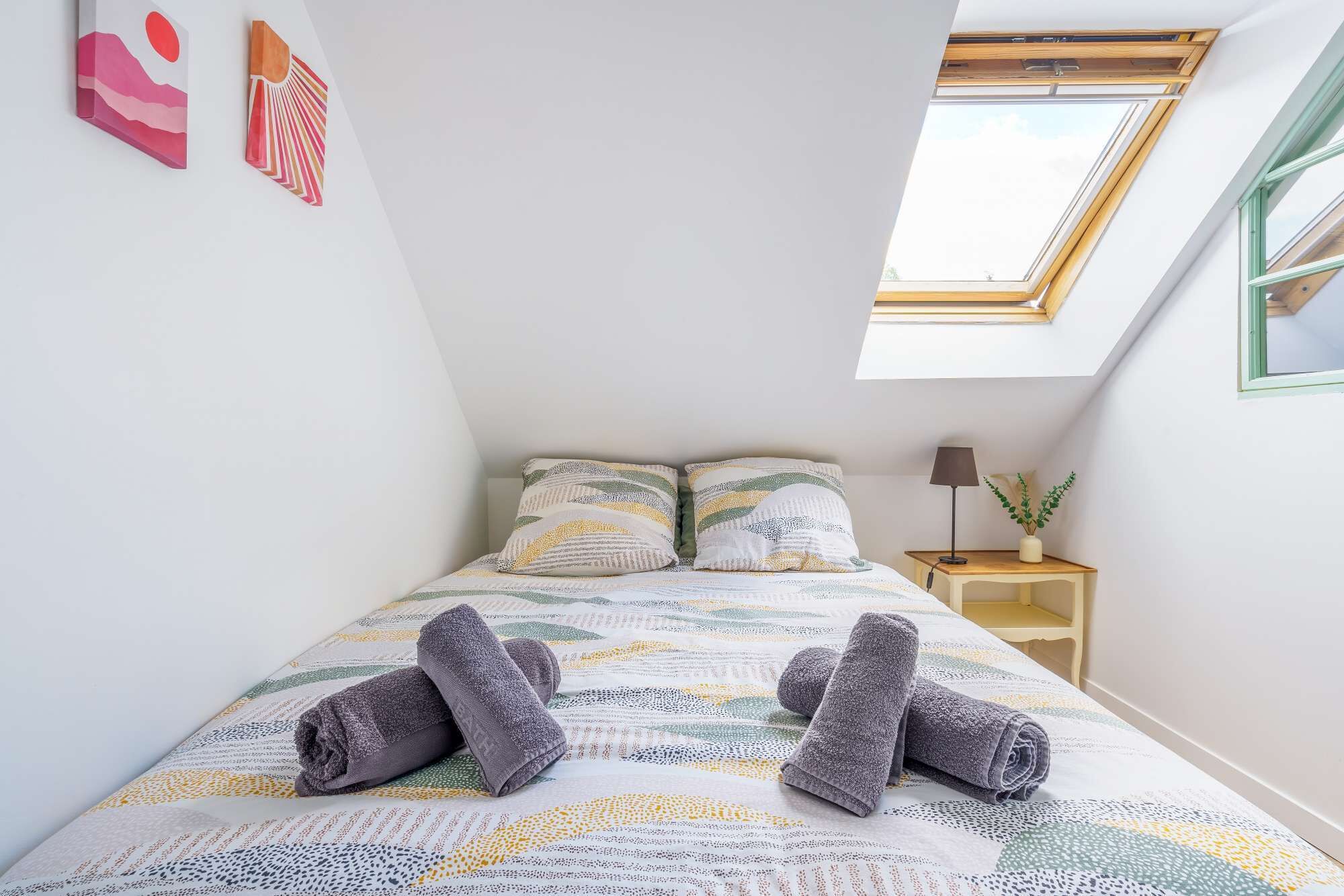 A bedroom with a skylight, a bed with patterned bedding, two pillows, a bedside table with a lamp, and a small plant.