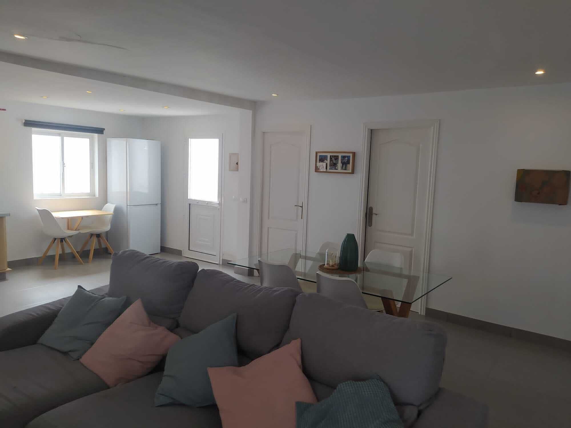 Ein modernes Wohnzimmer mit einem grauen Sofa, einem Glastisch und einem Essbereich mit weißem Tisch und Stühlen.