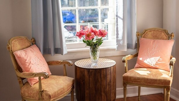Duas cadeiras ornamentadas com assentos e costas acolchoadas, dispostas em ambos os lados de uma mesa redonda de madeira com um vaso de vidro contendo flores cor-de-rosa.