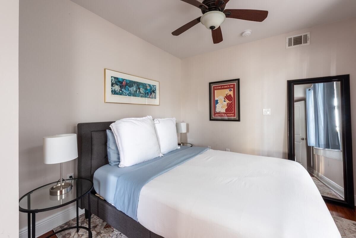A hotel room with a bed, a ceiling fan, a framed picture, a poster, and a window with curtains.