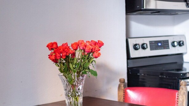 A wooden dining table with a vase of red flowers, a red upholstered chair, and a kitchen in the background. - Enchanting Garden District Magazine Street Flat (New Orleans)