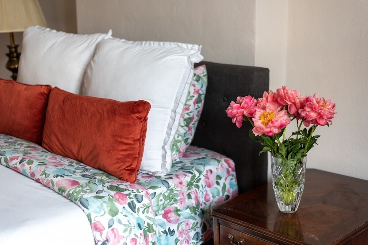 Ein ordentlich bezogenes Bett mit floraler Bettwäsche, eine Vase mit rosa Blumen und ein dunkler Nachttisch.