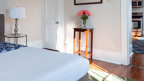 Un dormitorio con una cama, una mesita de noche con lámpara, una pequeña mesita auxiliar de madera con un jarrón de flores, un cuadro en la pared y una puerta abierta que da a una cocina.