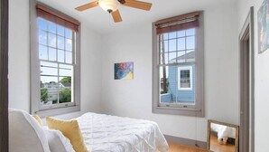 Een slaapkamer met een bed, twee ramen, een plafondventilator en een schilderij aan de muur.