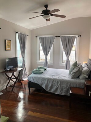 A bedroom with a bed, a desk with a TV, a ceiling fan, and two windows with curtains. - Delightful Freret St Studio Apartment w/parking (New Orleans)